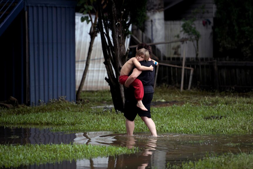 A woman carries a child into her property through ankle-deep water as floodwaters recede in Lismore.