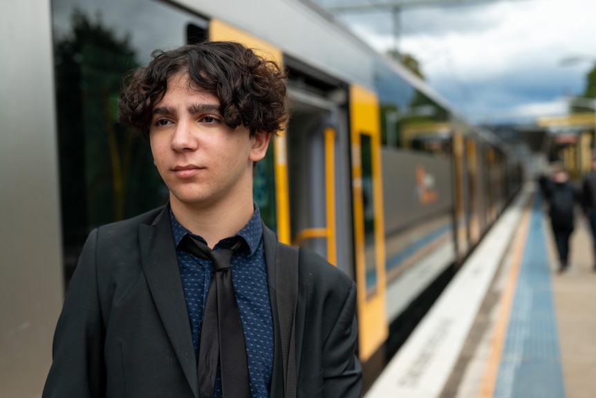 A young man wearing a suit and tie stands on a train station platform, a train is stopped next to him with its doors open.