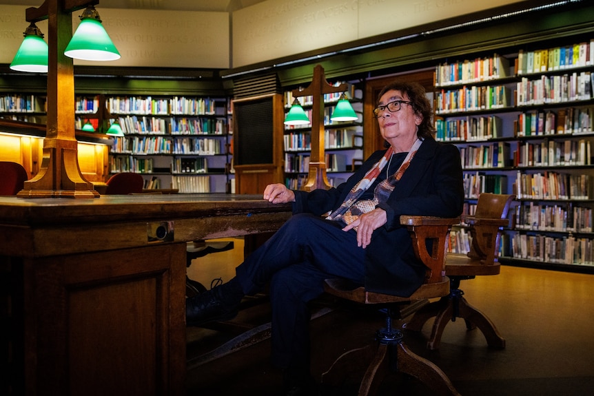 An older Indigenous woman with brown hair and glasses reclines in a chair in a book-lined library reading room