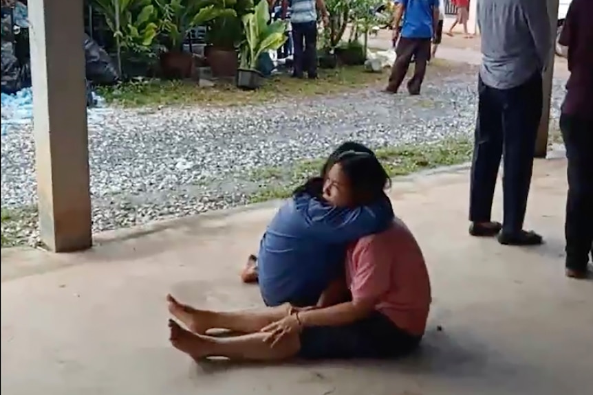 A distraught woman sits on the ground with her legs stretched out as she is comforted in a hug. 
