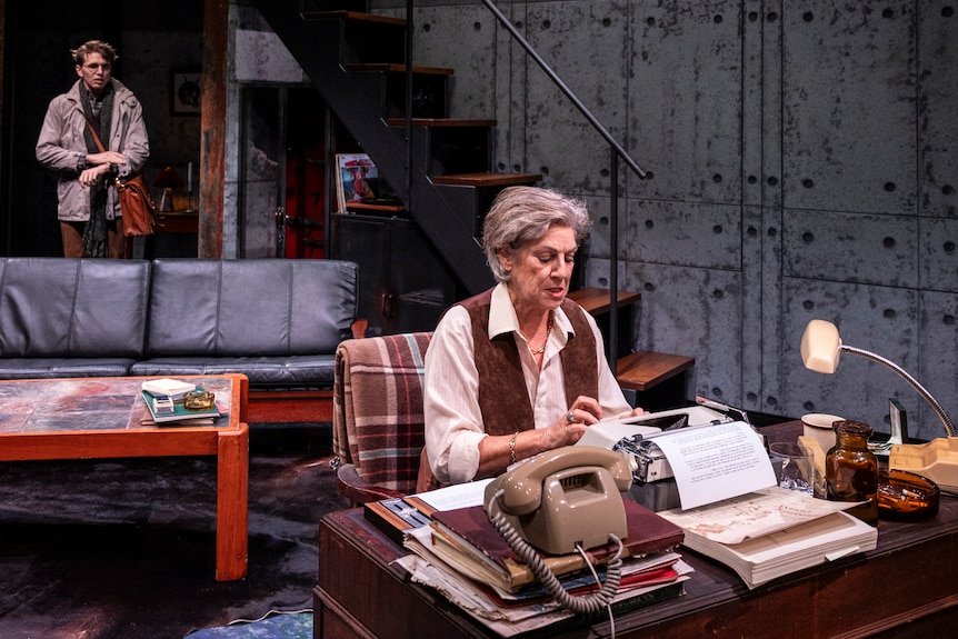On stage, Toni Scanlan sits at a desk, working on a typewriter. Behind her in a doorway stands an awkward Laurence Boxhall.