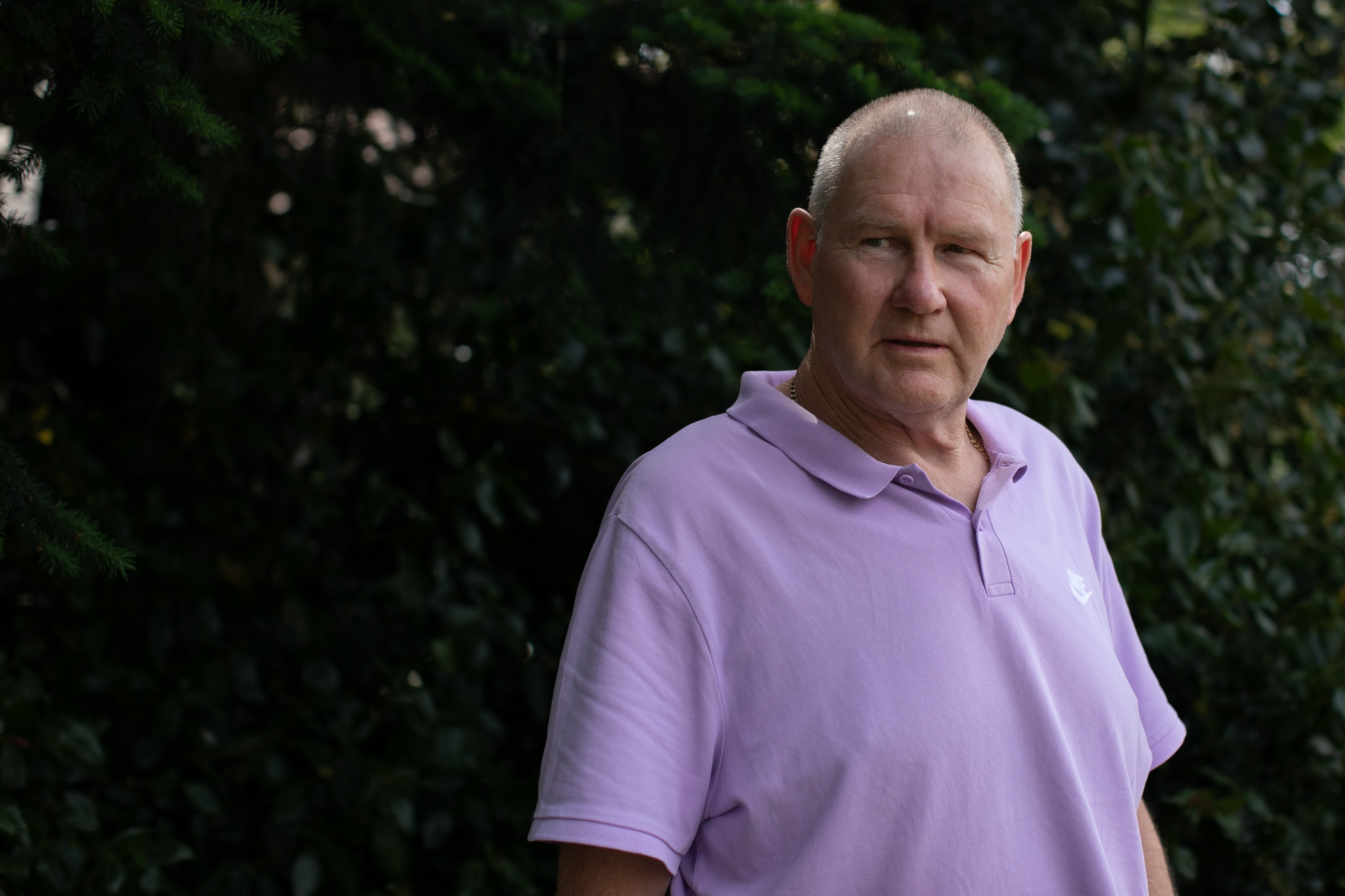 A man wearing lilac polo shirt stands in front of bushes, looking to left of camera
