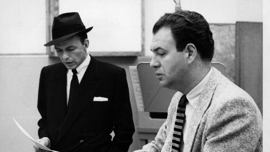 Frank Sinatra wearing a black suit and hat looking at a piece of paper. Nelson Riddle is sitting next to him in a grey suit.