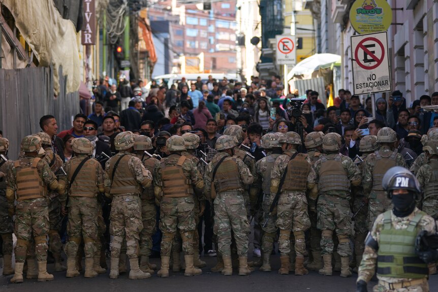 Several people in army gear block people walking towards them in a street.