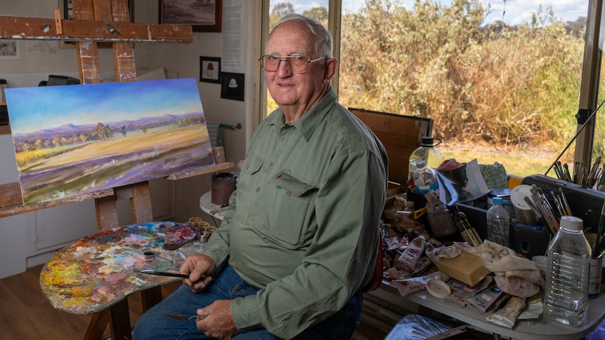 An older man sits in front of a canvas in an art studio.