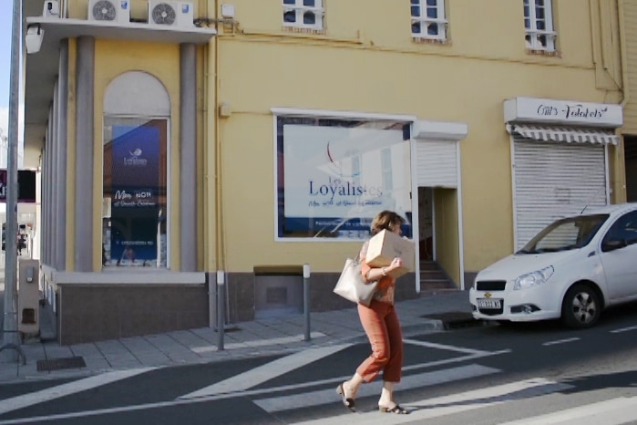 On a sunny day, you view a woman carrying a box uphill in front of a yellow French colonial-era building.