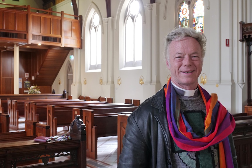 A man wearing a religious collar and a pride flag inside a church with stained-glass windows in the background