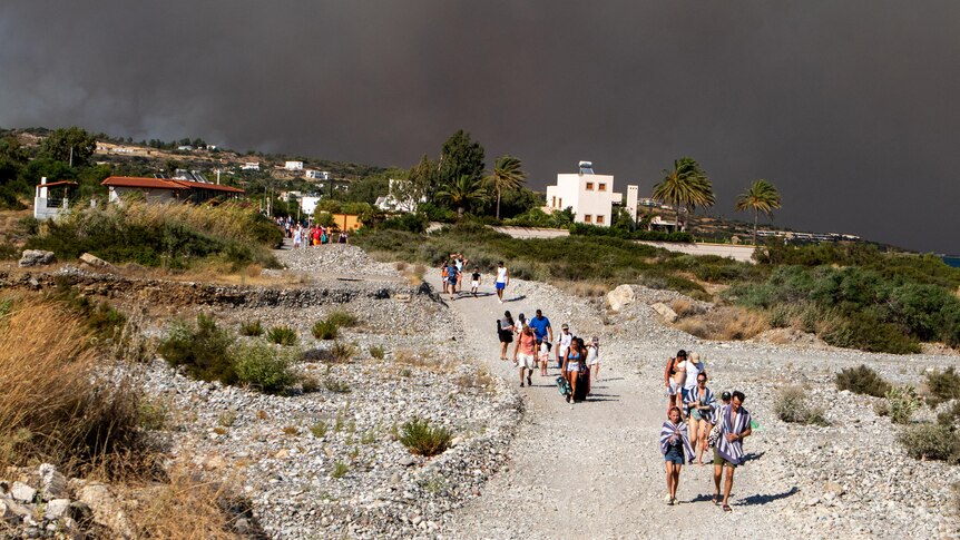 Tourists walk along a stony road in Rhodes as the sky behind them is filled with wildfire smoke