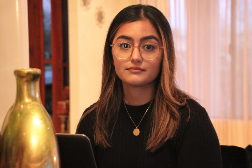 A woman with dark hair and glasses is sitting next to a gold vase with her laptop open