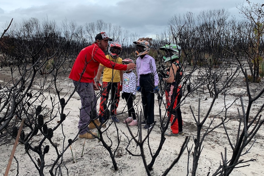 A man speaks to four people wearing motorcycle helmets among burnt-out trees