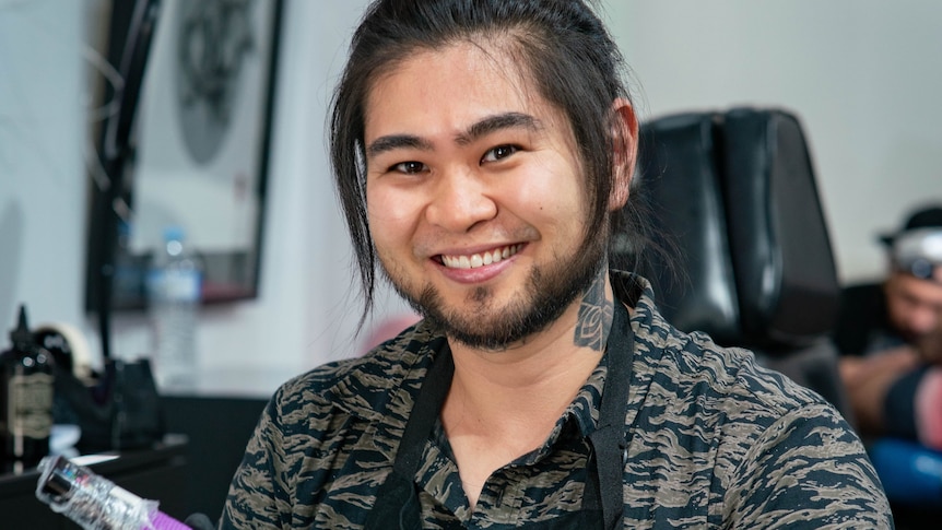 A man with Asian features is smiling, with a tattoo ink-injecting device in his hand. He's wearing a patterned shirt and apron