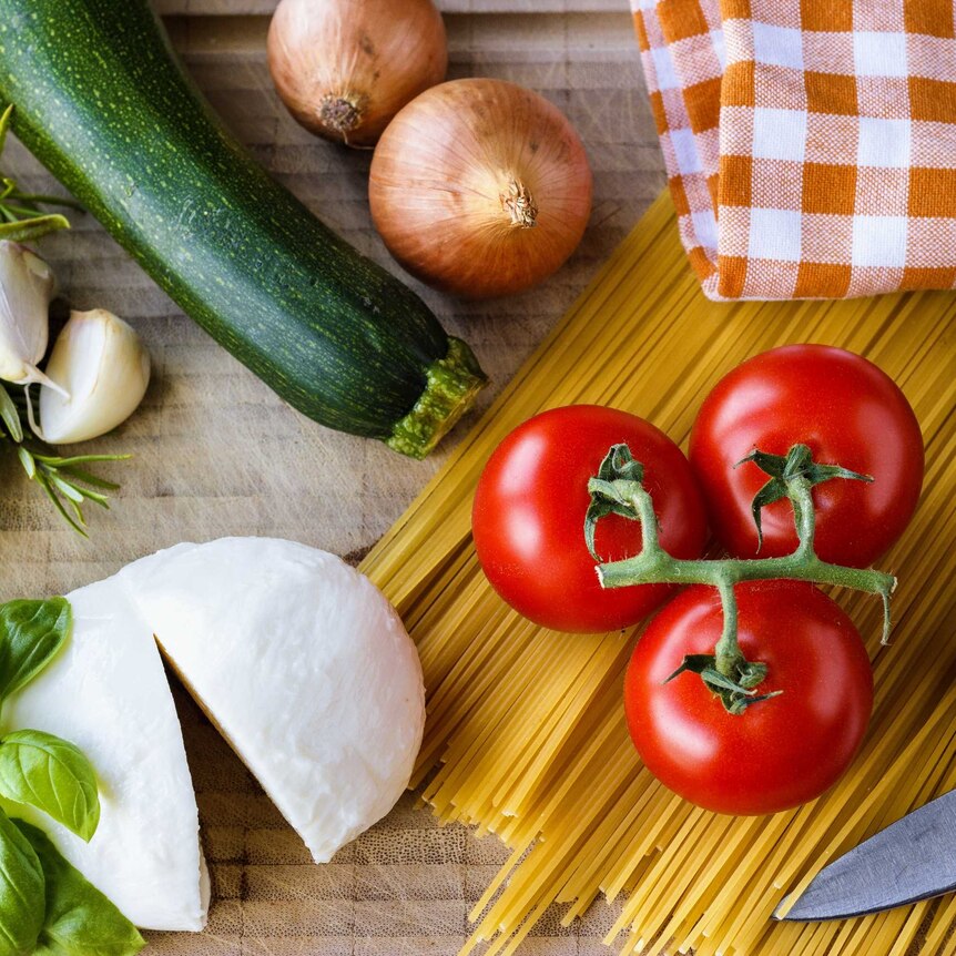 A flat-laid array of Italian ingredients including herbs, vine tomatoes, mozzarella, and pasta.