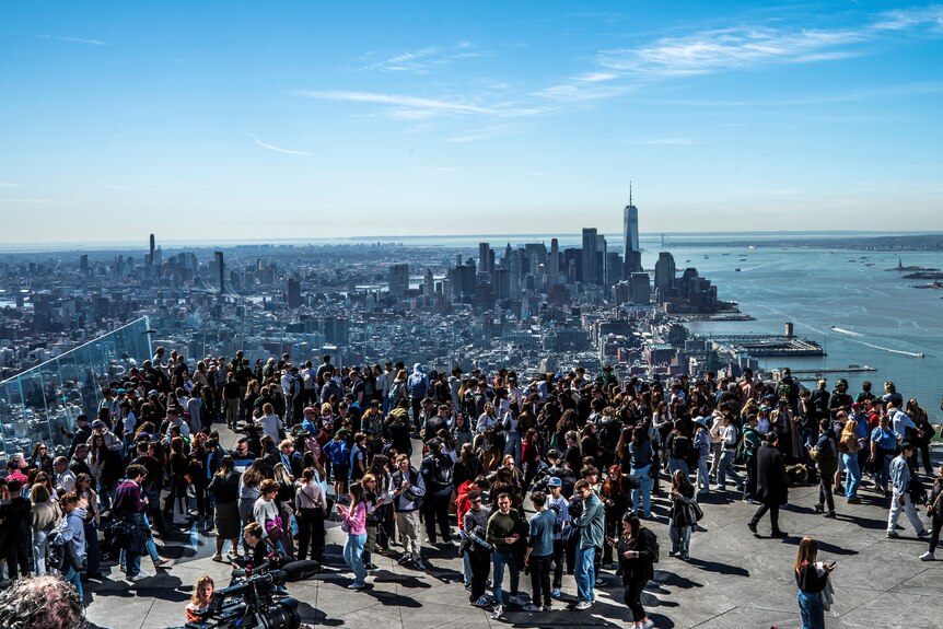 Dozens of people gather on a rooftop with a city skyline and bright blue sky in the background