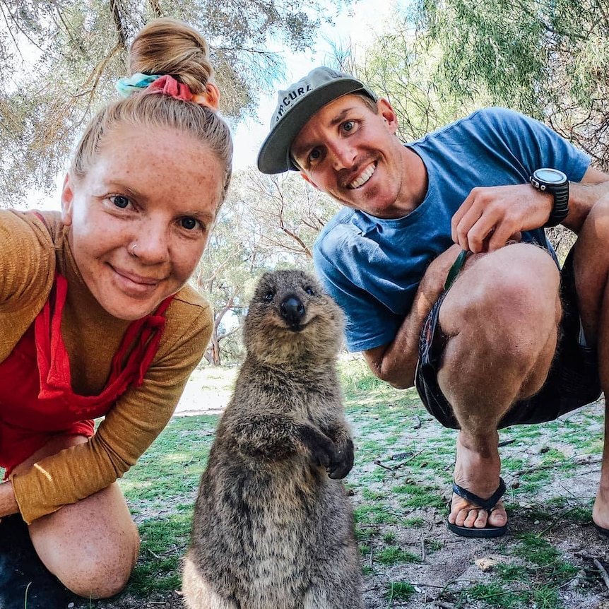 A quokka appears to be smiling at the camera while flanked by two people.