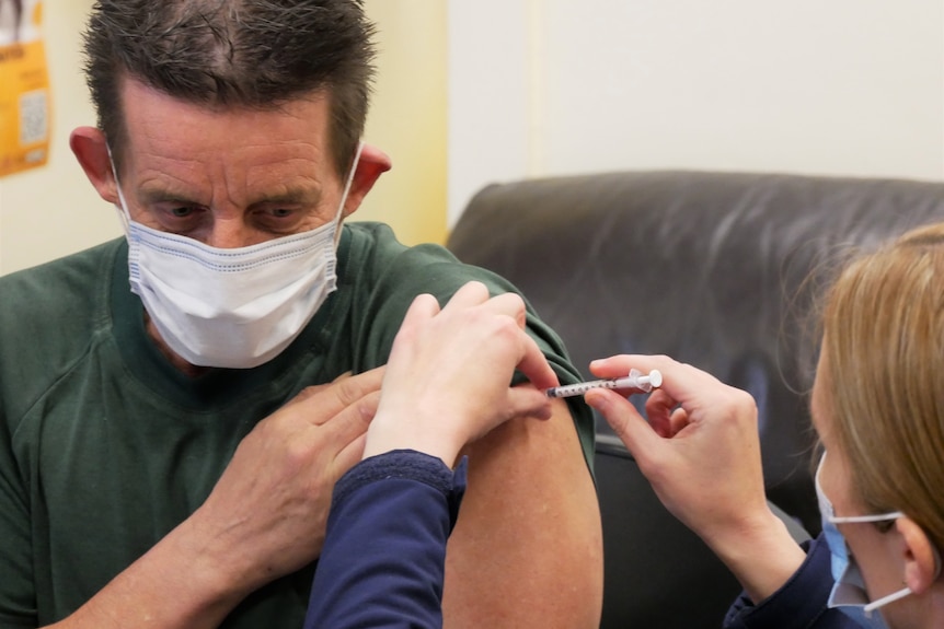 A man wearing a surgical mask holds his sleeve up and is injected with a needle by a nurse.
