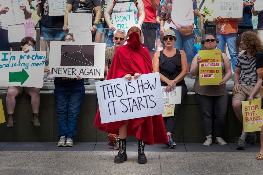 A woman in a Handmaid's Tale inspired outfit - red cloak and white bonnet - holds a sign reading 'This is How it Starts'