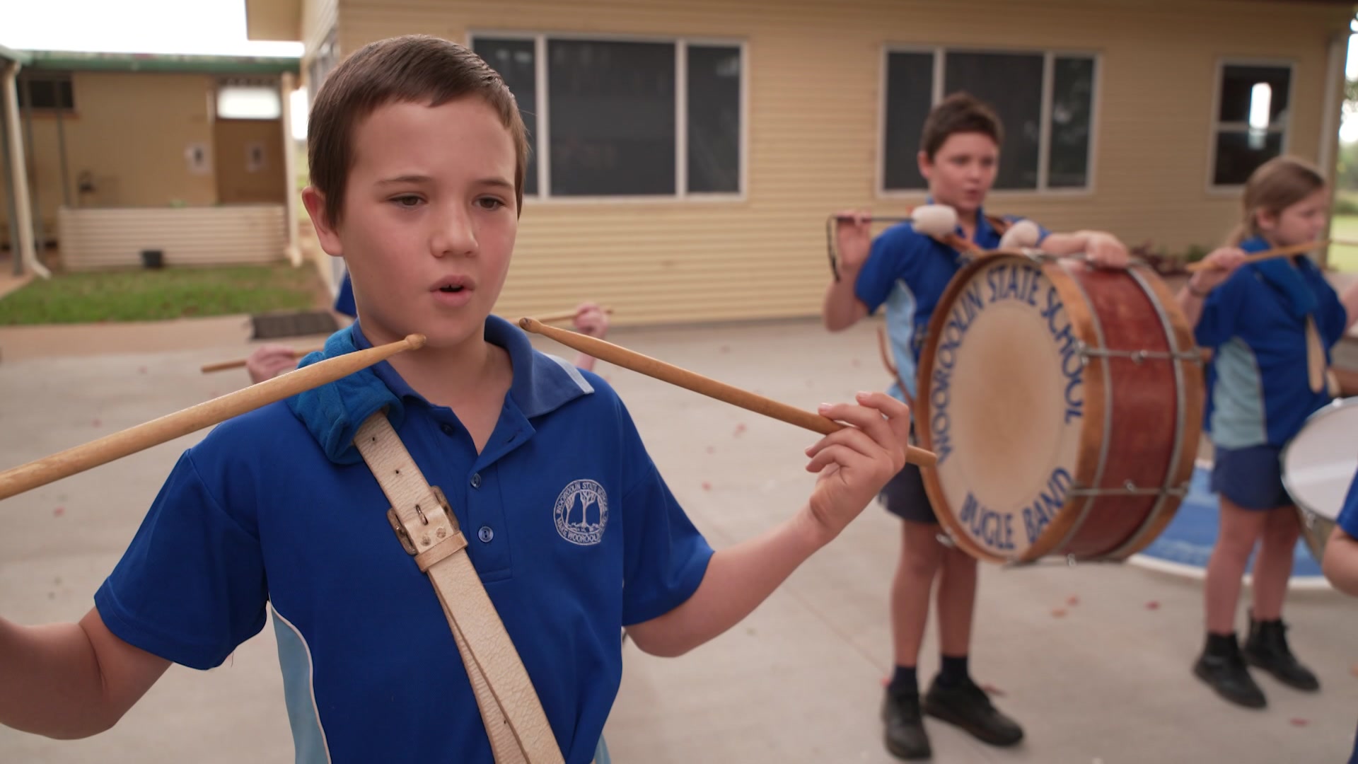 Young boy plays the drum in a marching band. He is wearing a blue school uniform.