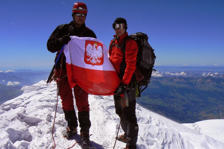 Two men stand atop of snowy mountain peak holding red and white flag