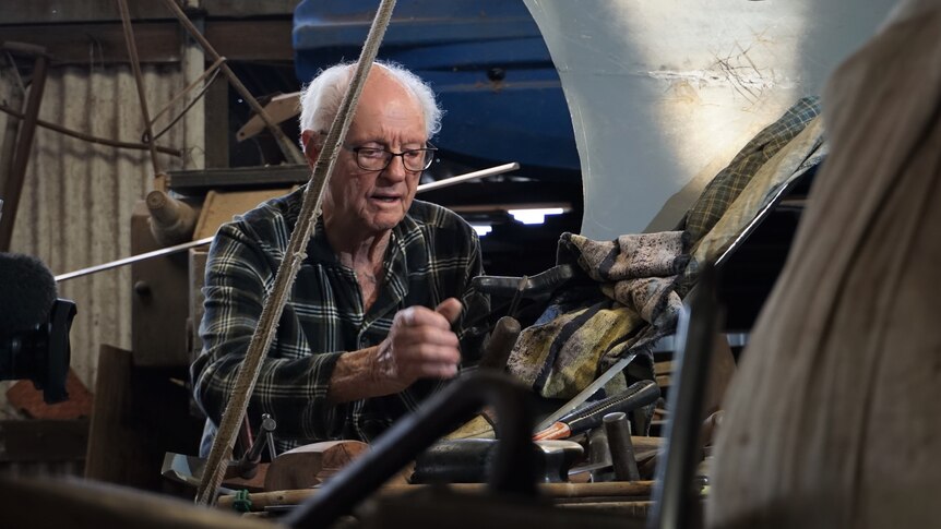 An old man wearing a flannel shirt tinkering with a car. 