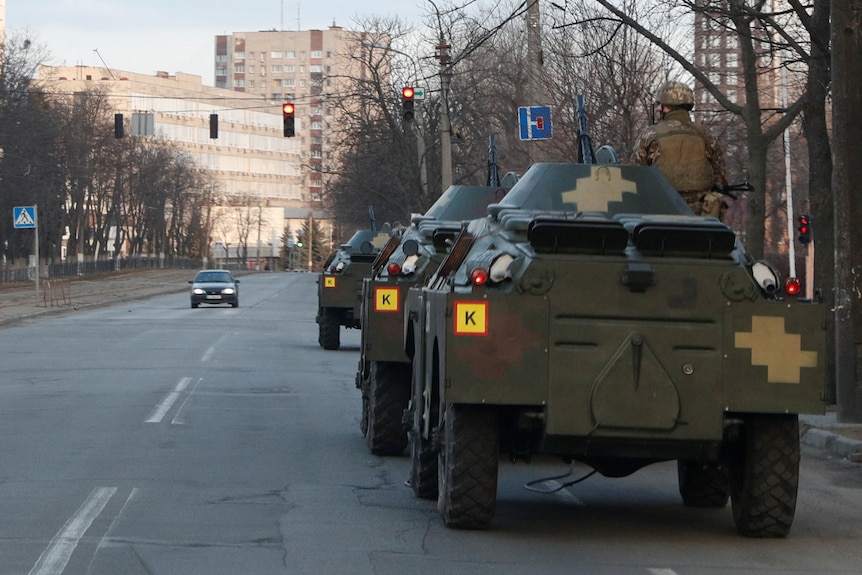 Three Ukrainian armoured personnel carriers parked on a road, with one serviceman sitting atop one of the vehicles.