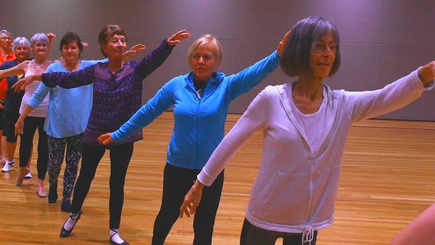A row of older womean in a ballet pose, left arms raised to the front, right arms lowered to the back