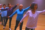 A row of older womean in a ballet pose, left arms raised to the front, right arms lowered to the back