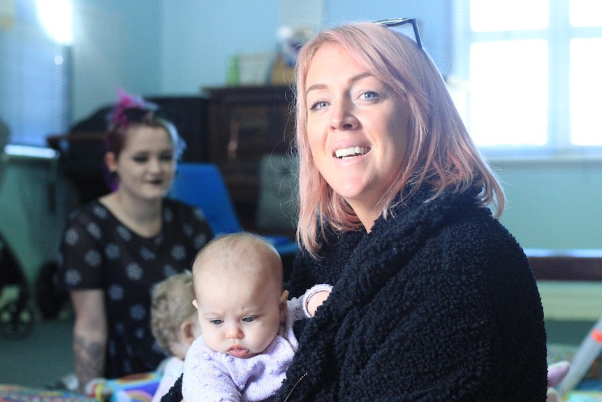 A woman with pink hair, holding a baby. A young woman is in the background