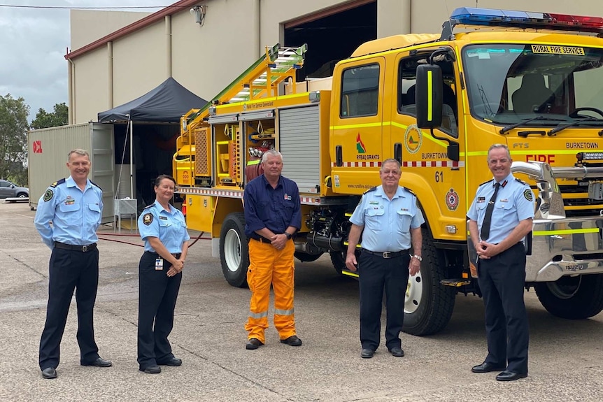 Four men and a woman, four of them in uniform, stand in front of a yellow fire truck on cloudy day.