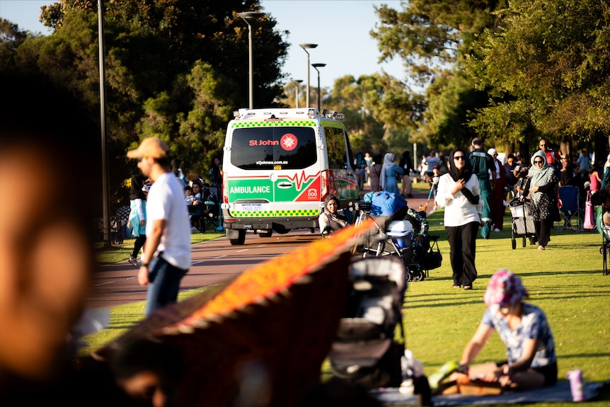 An ambulance travels on a road near a packed park in the afternoon.