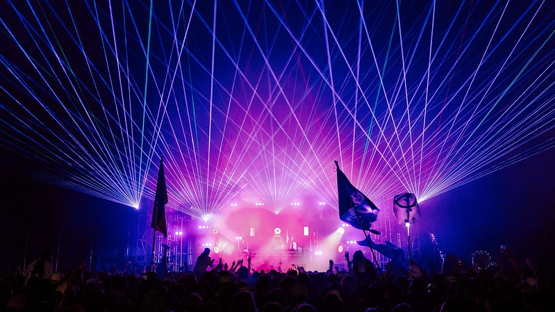Night photo of a festival crowd with pink lighting and blue lasers fanning out from the stage