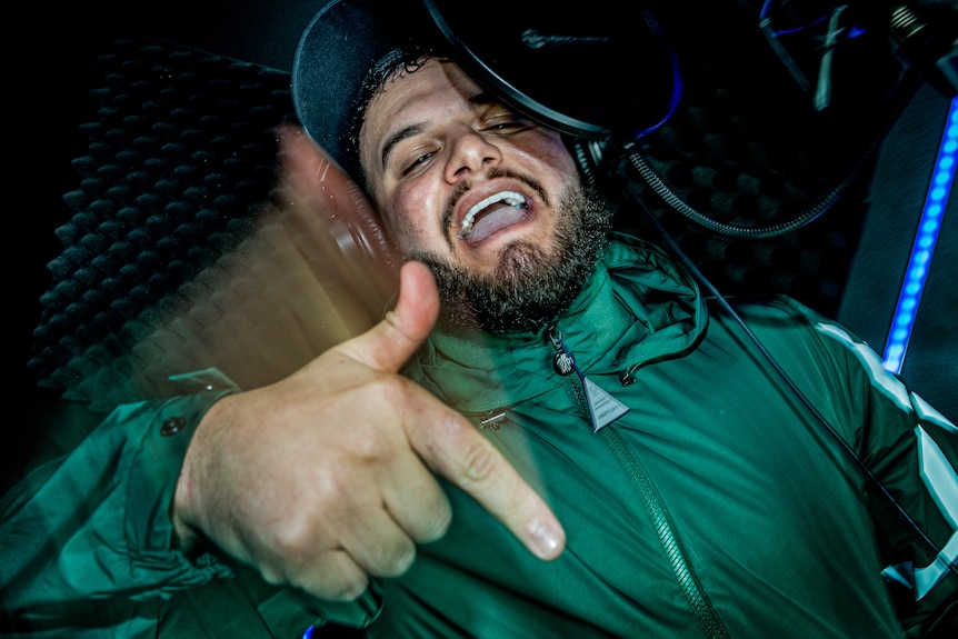 A man in a recording booth, wearing headphones, raps into a microphone. He's making a pointing gesture moving his hand down.