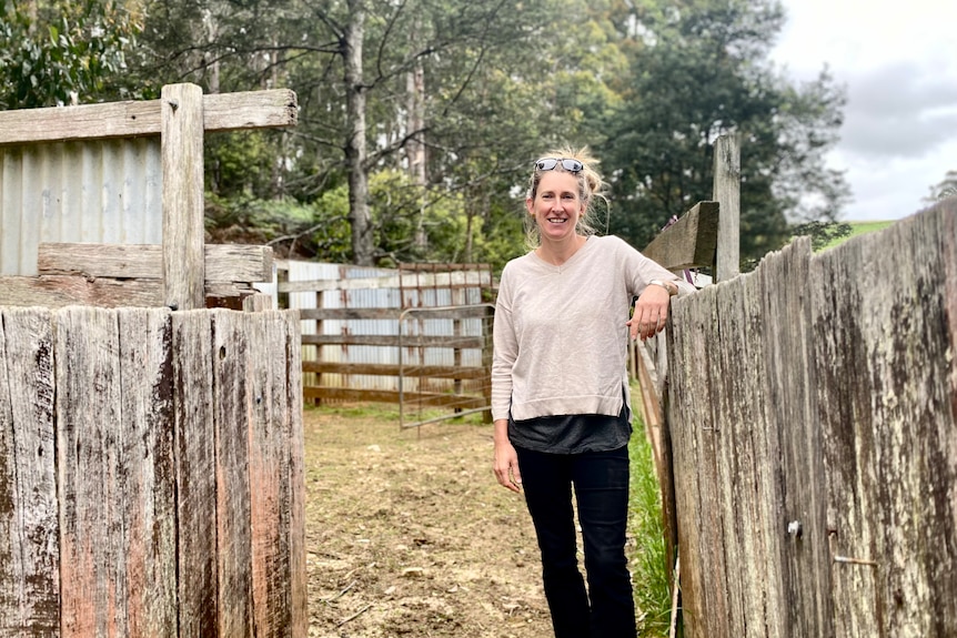 A smiling woman in work gear leans on an old fence at a farm.