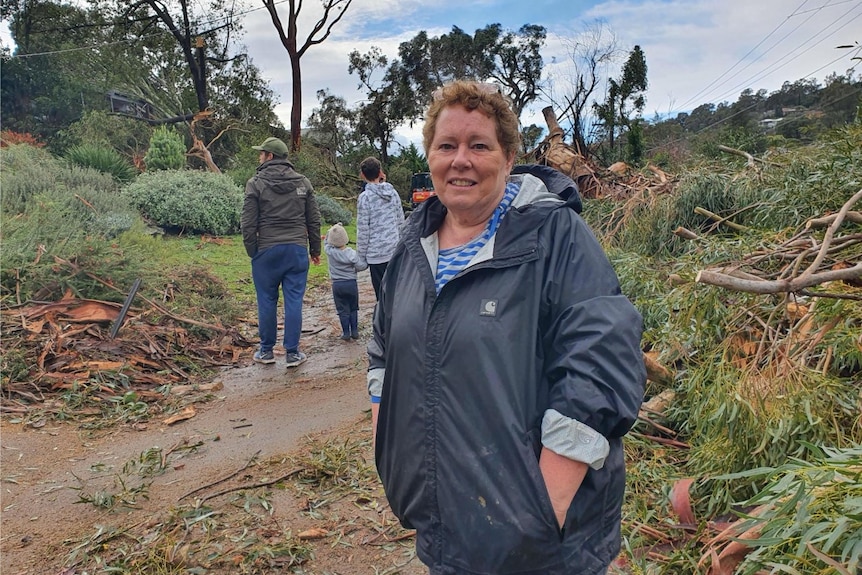 Carolyn stands, dressed in a jacket, on a street strewn with fallen trees and debris.