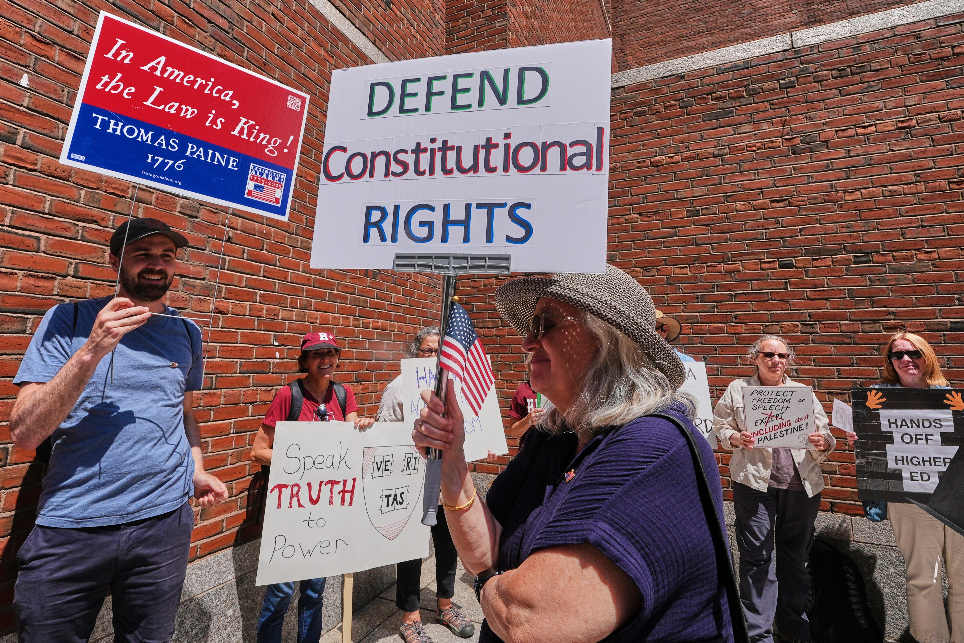 A group of people hold protest signs in front of a brick building. One says defend constitutional rights. 