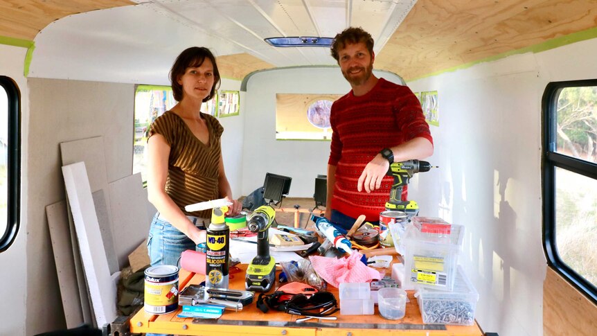 A female and male stand in their bus which is being renovated, with power tools on a table in front of them.