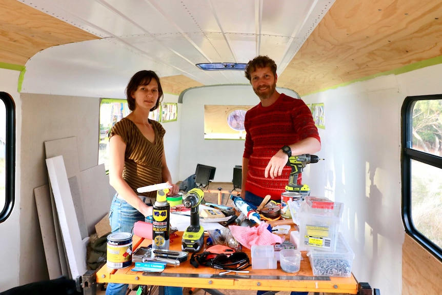 A female and male stand in their bus which is being renovated, with power tools on a table in front of them.