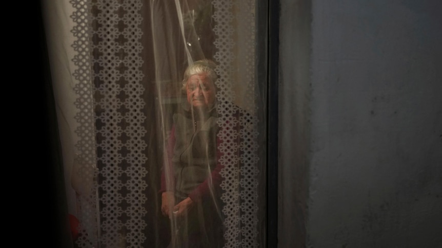 A woman sits in a room on a makeshift bed. She is veiled by a curtain hanging across a doorway.