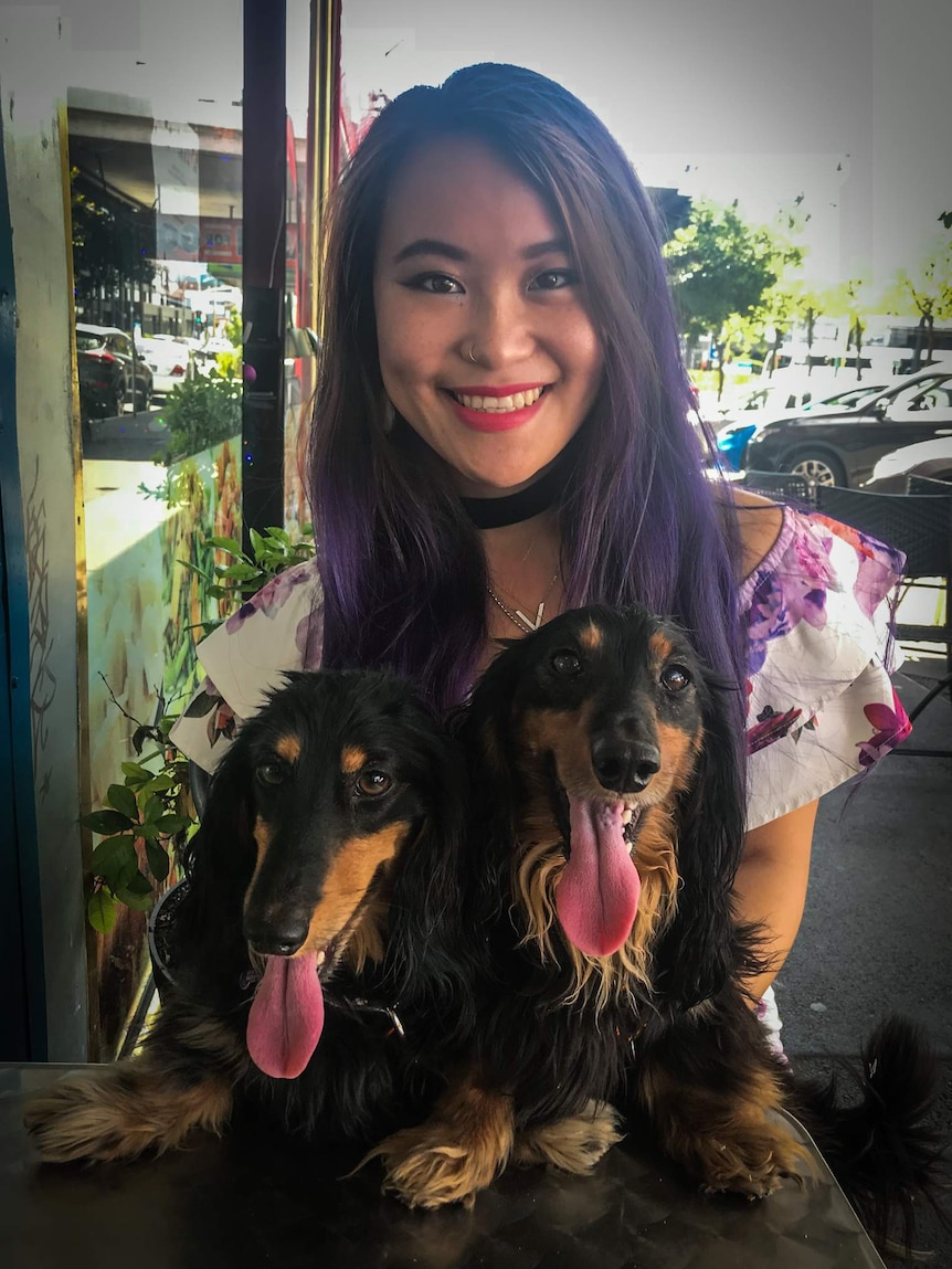 A young woman wearing a floral dress sits at an outdoor cafe table holding two dogs on her lap.