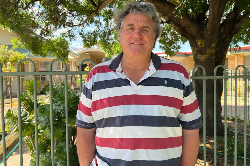 A man with grey salt and pepper hair wearing a blue, white and red striped polo shirt in front of a green gate