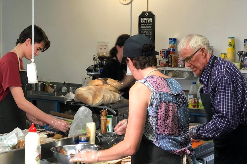 A woman, man and teenage boy wear aprons and gloves to make sandwiches in a kitchen.