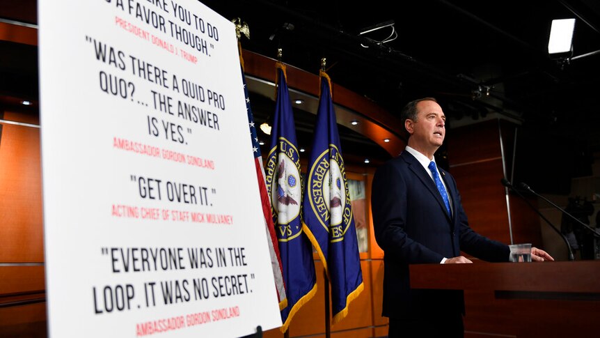 A large white placard is mounted on a black easel as Adam Schiff stands behind a wooden lectern.