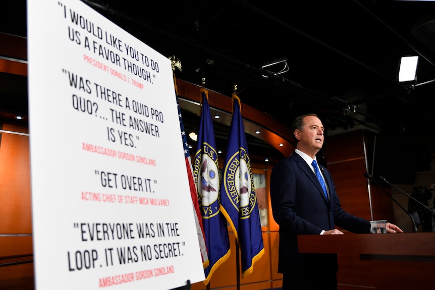 A large white placard is mounted on a black easel as Adam Schiff stands behind a wooden lectern.