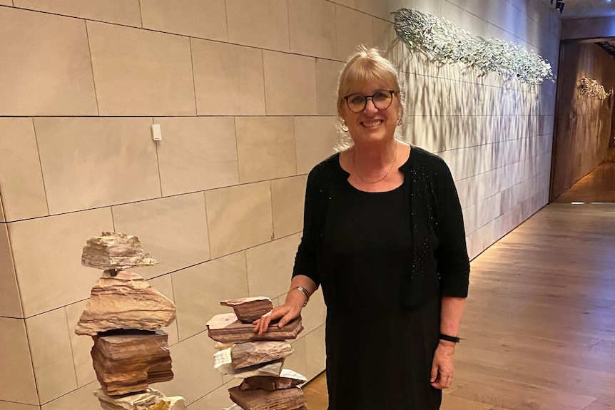 A woman in a black dress and glasses stands next to a sculpture.