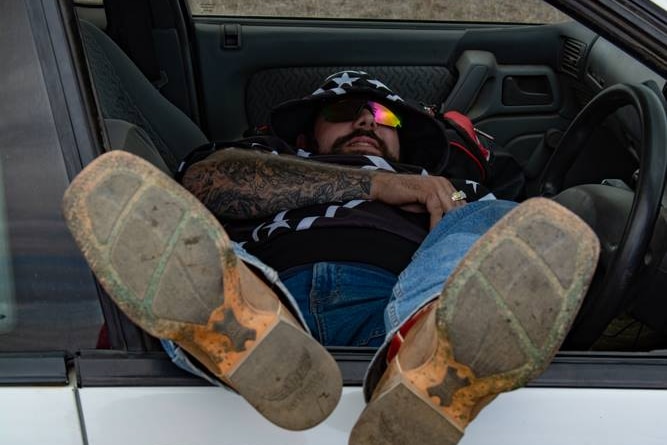 A tattooed man with dark sunglasses and a bandana lies across the front seats of his car with his feet out the driver's window