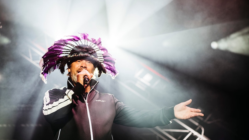 Jay Kay of Jamiroquai stands on stage singing into a microphone with his arm outstretched. He wears a feathered headdress.