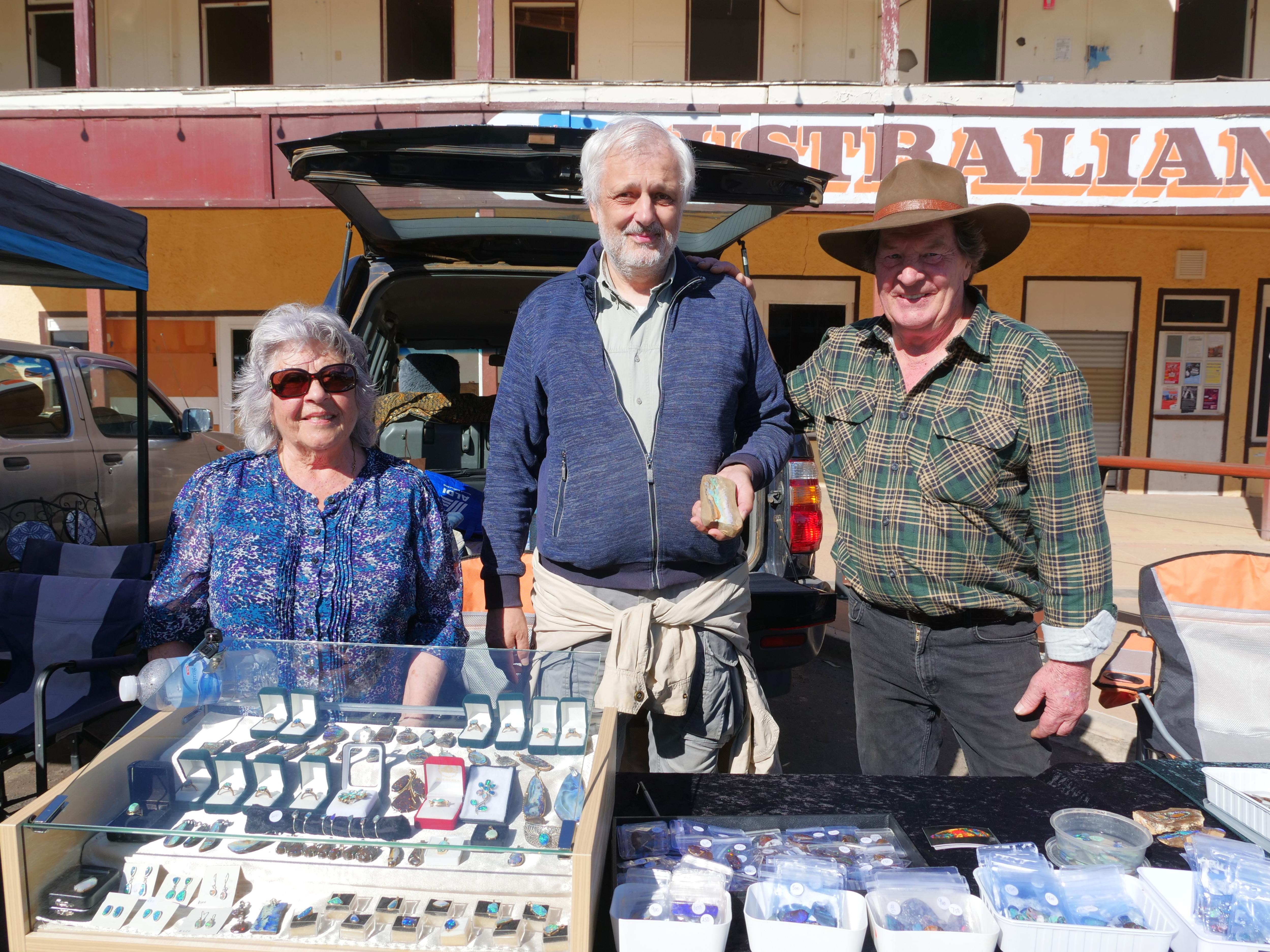 Three smiling older people behind a stall at an open-air market.