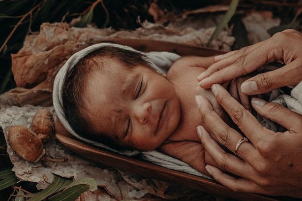 A newborn baby sleeps in a wooden cradle among paperbark and leaves. 