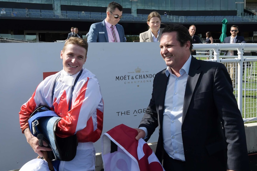 A jockey carries his saddle as the trainer smiles in the mounting yard after winning a race.