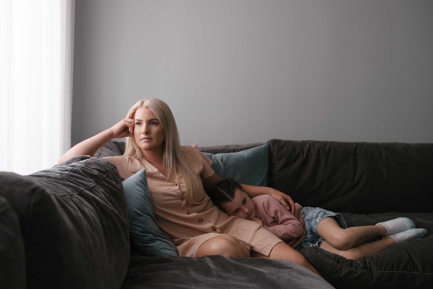 A blonde woman looks out the window from her couch with her daughter lying on her lap