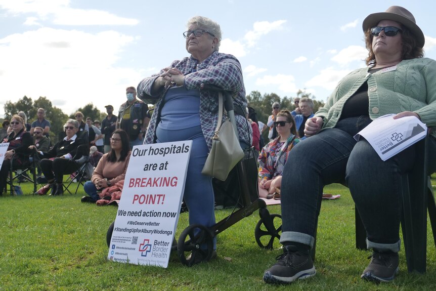 A woman sits in a wheelchair with a sign at her feet pushing for a new hospital. 
